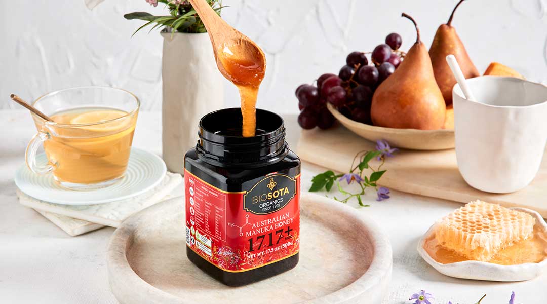 Biosota Manuka honey MGO 1717+ jar with honey dripping from a wooden spoon, beside a cup of lemon tea